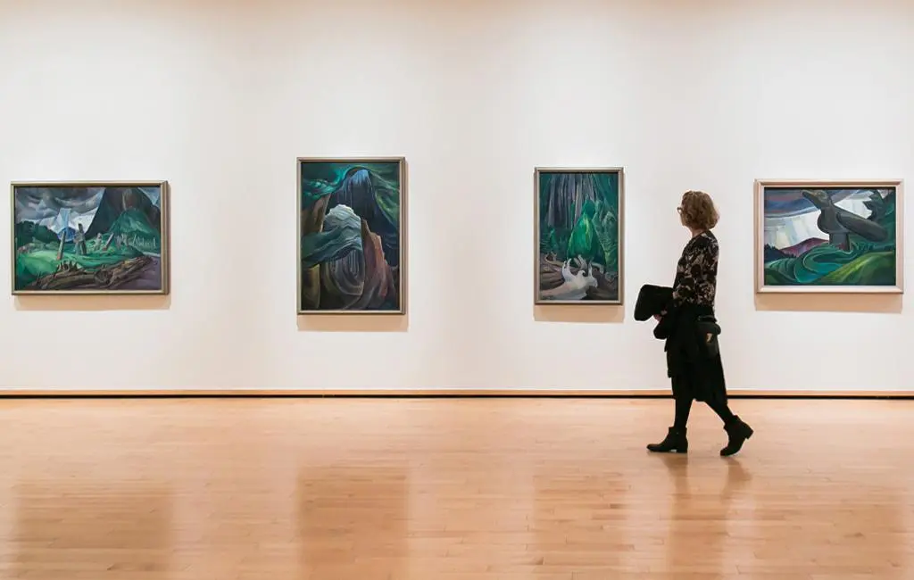 Woman walking in from of four Emily Carr paintings of the forest.